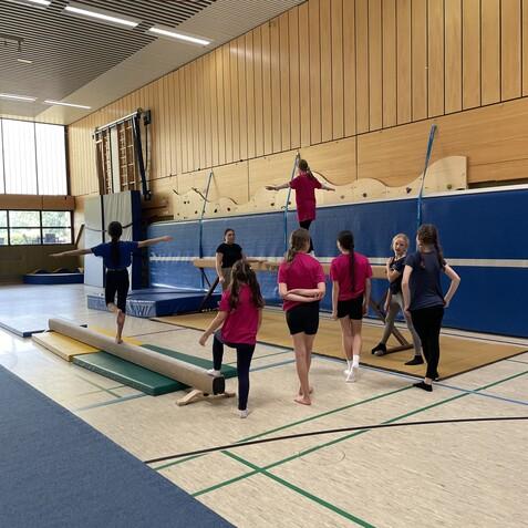 Sechs Kinder üben auf einem Balancierbalken in einer Sporthalle. Eine Trainerin beobachtet und gibt Anweisungen.