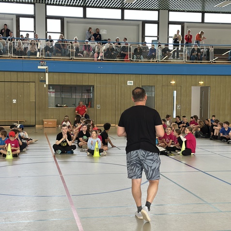 Lehrer spricht vor versammeltem Publikum von Kindern in Sportkleidung in einer Schulturnhalle. Zuschauer aus erh&ouml;hter Position.
