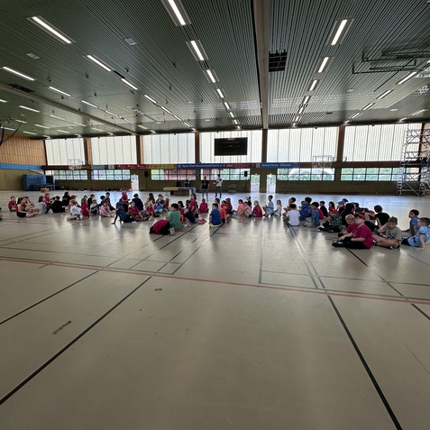 Gruppensitzung von Kindern in einer Sporthalle, farbenfrohe Kleidung, mit Gymnastikmatte und gro&szlig;en Fenstern im Hintergrund.