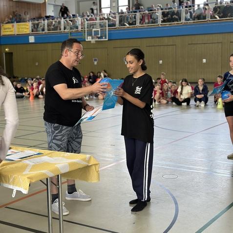 Ein Mann &uuml;berreicht einer jungen Frau ein Zertifikat in einer Sporthalle, umgeben von Zuschauern auf Trib&uuml;nen.
