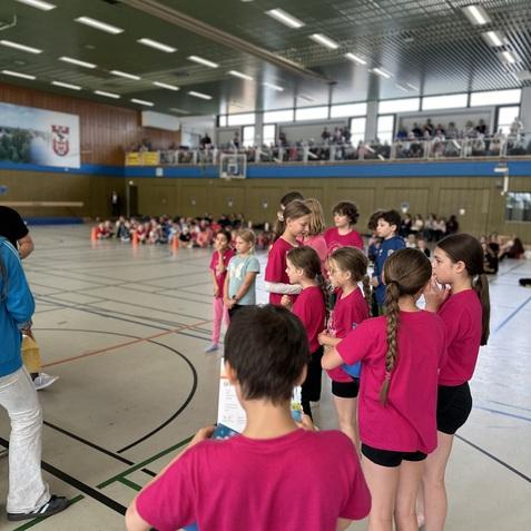 Gruppe von Kindern in pinken T-Shirts steht in einer Sporthalle, w&auml;hrend eine Person Anweisungen gibt.