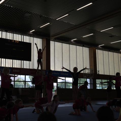 Gruppe von Kindern in Sportkleidung bei einem Turntraining, einige stehen auf H&auml;nden anderer, mit Wanduhr im Hintergrund.