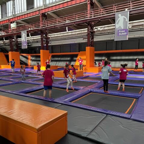 Kinder in bunten T-Shirts springen auf Trampolinen in einer Indoor-Trampolinhalle mit orangenen und lilafarbenen Elementen.