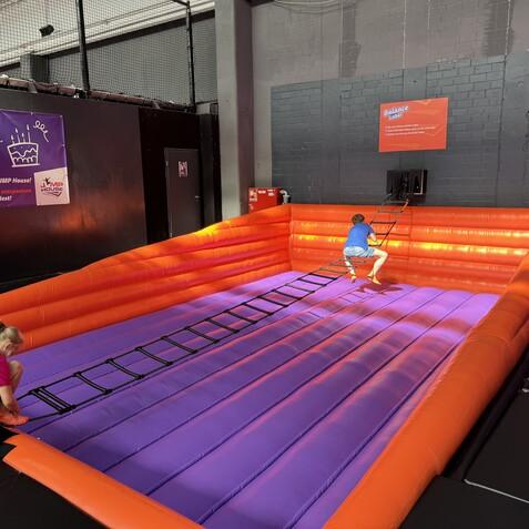 Zwei Kinder spielen auf einer h&uuml;pfenden, orange-lila H&uuml;pfburg mit einer Leiter im JUMP House.