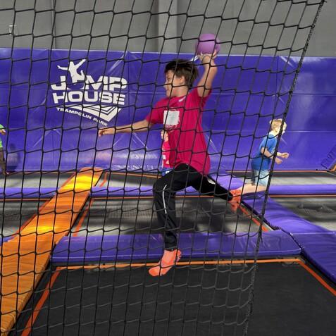 Junge in pinkem Shirt springt mit Ball in einem Trampolinpark, Hintergrund mit Netzen und anderen Kindern.