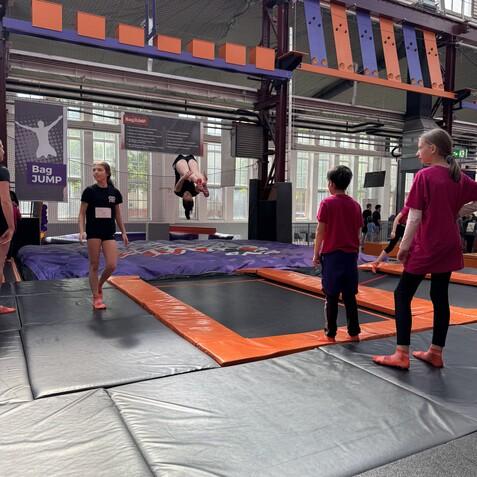 Eine Person springt auf einem Trampolin, w&auml;hrend andere Zuschauer in Sportsachen warten. Helle Halle mit gro&szlig;en Fenstern.
