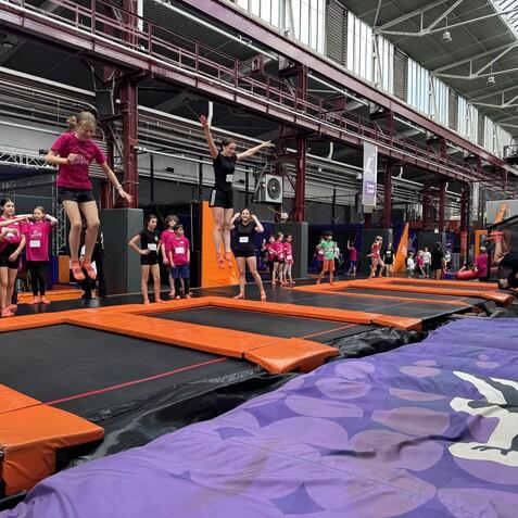 Gruppe von Kindern in pinken T-Shirts springt auf Trampolinen in einer offenen Halle mit orangefarbenen Aufprallfl&auml;chen.