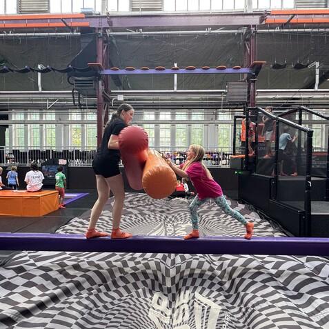 Zwei Kinder auf einer Schwebebalken-ähnlichen Plattform, kämpfen spielerisch mit großen, weichen Ballen in einer Trampolinhalle.
