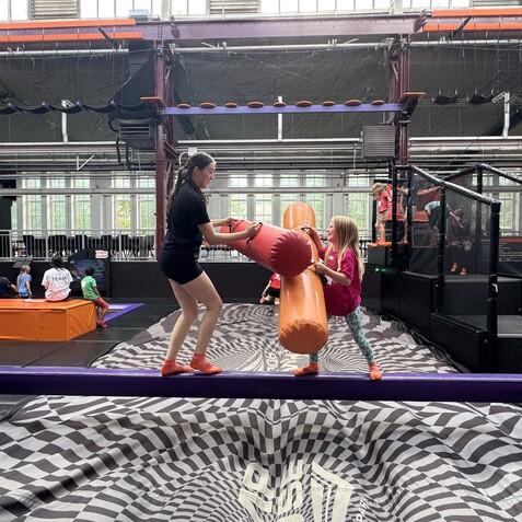 Zwei Kinder kämpfen spielerisch auf einem Balancebalken in einer Trampolinhalle. Im Hintergrund sind weitere Kinder aktiv.