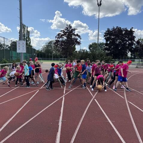 Gruppe von Kindern in bunten T-Shirts auf einer Laufbahn, einige halten Bälle, während sie sich zur Aktivität versammeln.