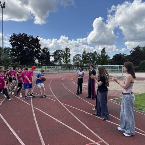 Gruppenspiel von Kindern auf einem Sportplatz, während einige Erwachsene am Rand applaudieren. Wolkiger Himmel im Hintergrund.