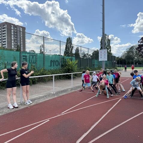 Zwei Trainerinnen beobachten eine Gruppe von Kindern, die sich auf einem Sportplatz zum Wettlauf vorbereiten.