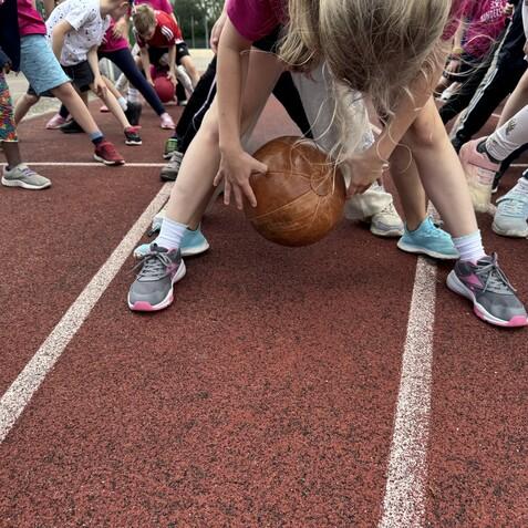 Kinder mit verschiedenen Sportschuhen beugen sich über einen Basketball auf einer roten Laufbahn.