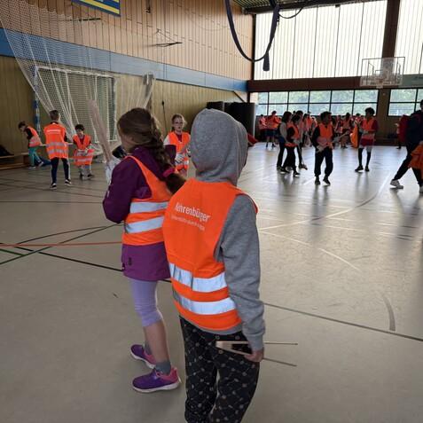 Kinder in orangefarbenen Warnwesten spielen in einer Sporthalle mit unterschiedlichen Aktivitäten und Materialien.