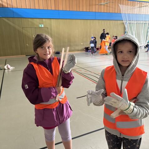 Zwei Kinder in Warnwesten halten Holzstöcke in einer Sporthalle voller Spiele und Aktivitäten.