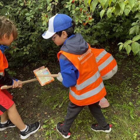 Zwei Kinder in Warnwesten betrachten eine Karte und halten eine Zange unter einem dichten Busch mit Beeren.