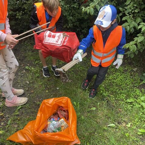 Kinder im Spiel angezogen mit Warnwesten sammeln Müll mit einer roten Tasche und einem orangefarbenen Müllsack.