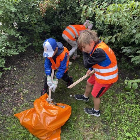 Drei Kinder in roten Warnwesten sammeln Müll in einem grünen Gebüsch und füllen ihn in einen orangefarbenen Sack.
