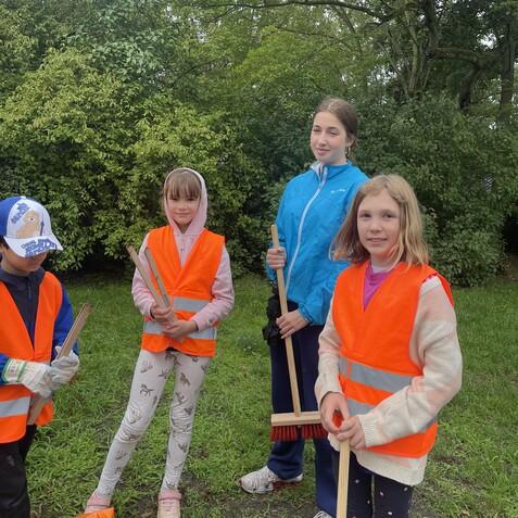 Vier Kinder in leuchtend orangefarbigen Westen stehen im Freien, bereit mit Besen und M&uuml;lls&auml;cken zu arbeiten.