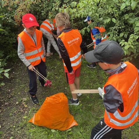 Gruppe von Kindern in orangenen Warnwesten sammelt M&uuml;ll in einem Gr&uuml;nbereich, umgeben von Str&auml;uchern und B&auml;umen.