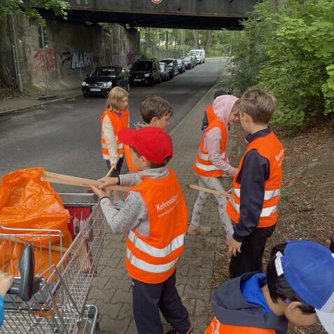 Gruppe von Kindern mit Warnwesten, die an einer Straße neben einem Einkaufswagen mit Müllsäcken arbeiten.