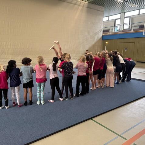 Eine Gruppe von Kindern &uuml;bt in einer Turnhalle, w&auml;hrend ein M&auml;dchen einen Sprung ausf&uuml;hrt.
