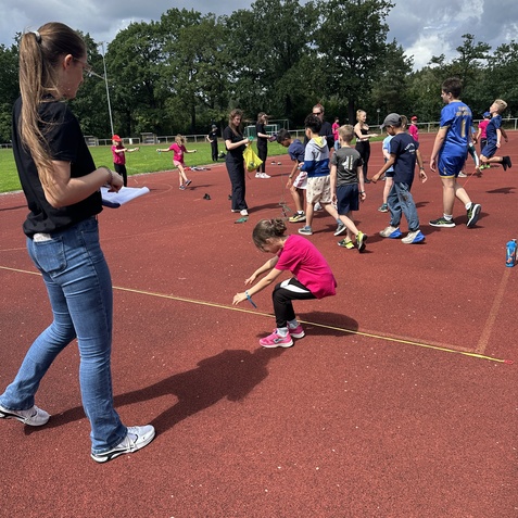 Kinder in Sportkleidung &uuml;ben auf einem L&auml;uferfeld, w&auml;hrend eine Trainerin Notizen macht. Wolkiger Himmel im Hintergrund.