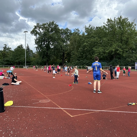 Gruppenspiel auf einem Sportplatz mit Kindern, die in bunter Sportkleidung aktiv sind, im Hintergrund gr&uuml;ne B&auml;ume.