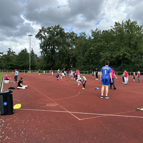 Gruppen von Kindern und Jugendlichen spielen auf einem roten Sportplatz, umgeben von B&auml;umen und Wolken.