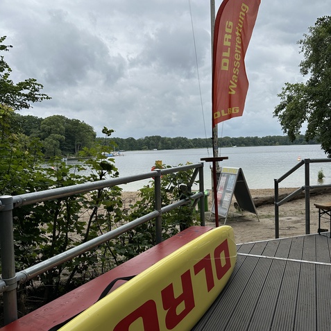 Blick auf einen See mit DLRG-Wasserrettungsflagge und einem Rettungsbrett auf der Holzterrasse. Wolkiger Himmel im Hintergrund.