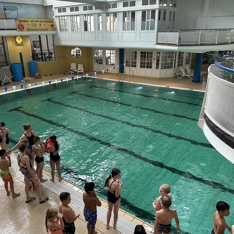 Kinder stehen in Badebekleidung am Beckenrand eines Schwimmbeckens mit Wassertiefe, das von einem Hallenraum umgeben ist.