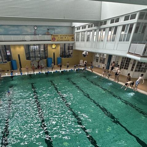 Schwimmbad mit mehreren Kindern, die im Wasser spielen, und einigen Erwachsenen am Beckenrand. Hell erleuchtet.