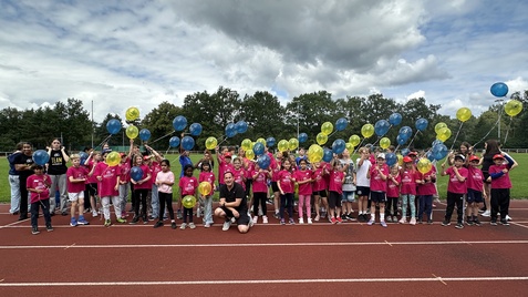 Gruppe von Kindern in pinken T-Shirts mit bunten Ballons auf einer Laufbahn, unter wolkigem Himmel.