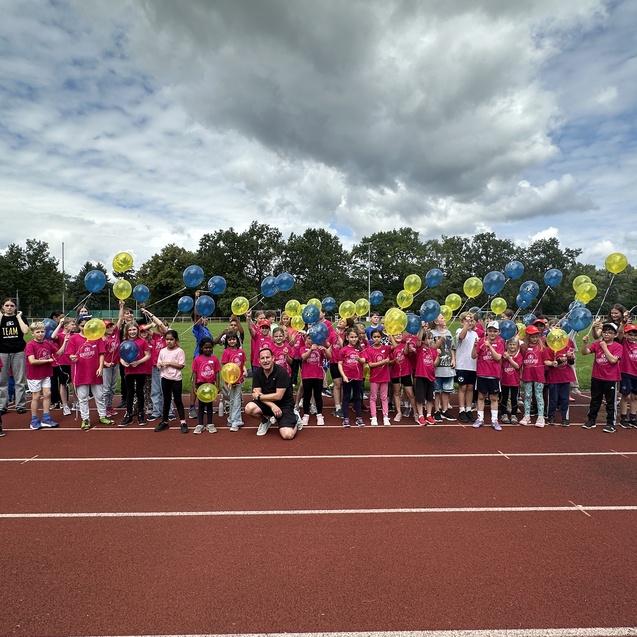Gruppe von Kindern in pinken T-Shirts mit bunten Ballons auf einer Laufbahn, unter wolkigem Himmel.