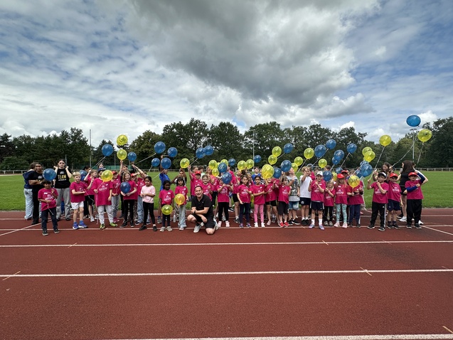 Gruppe von Kindern in pinken T-Shirts mit bunten Ballons auf einer Laufbahn, unter wolkigem Himmel.