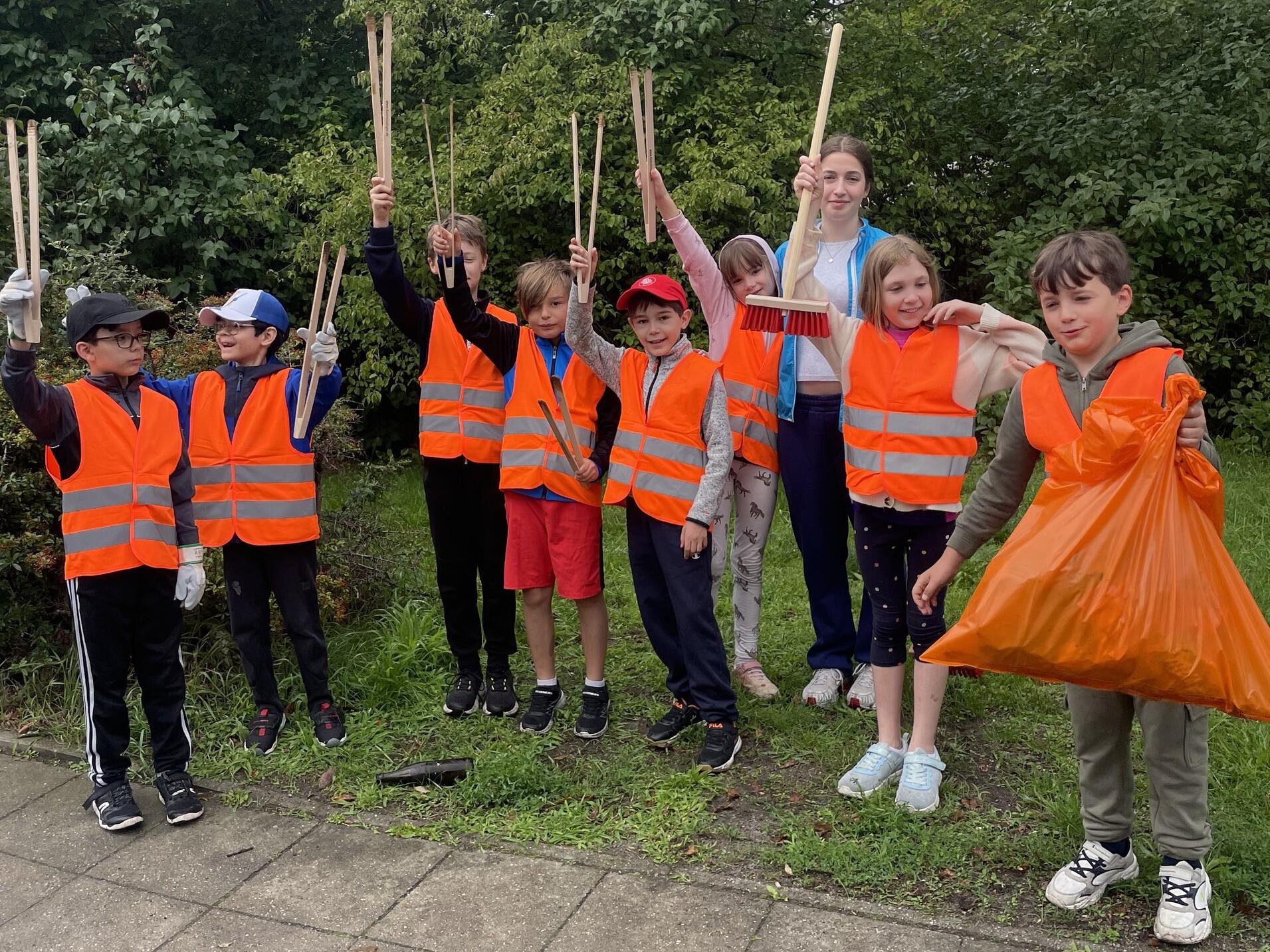 Gruppe von Kindern in orangefarbenen Warnwesten mit Müllzangen und einem Müllsack, stehend in einer Grünanlage.