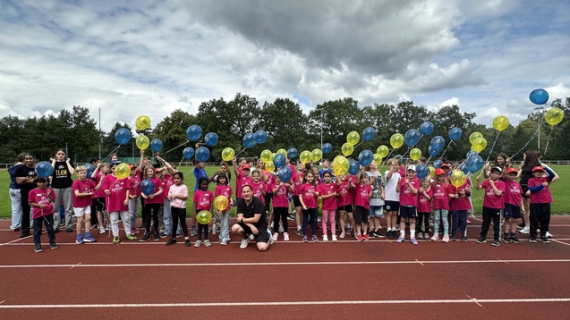 Gruppe von Kindern in pinken T-Shirts mit bunten Ballons auf einer Laufbahn, unter wolkigem Himmel.