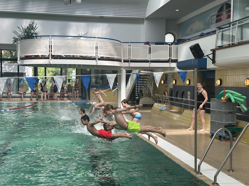 F&uuml;nf Kinder springen ins Wasser eines Schwimmbeckens, w&auml;hrend eine Trainerin am Beckenrand zusieht.