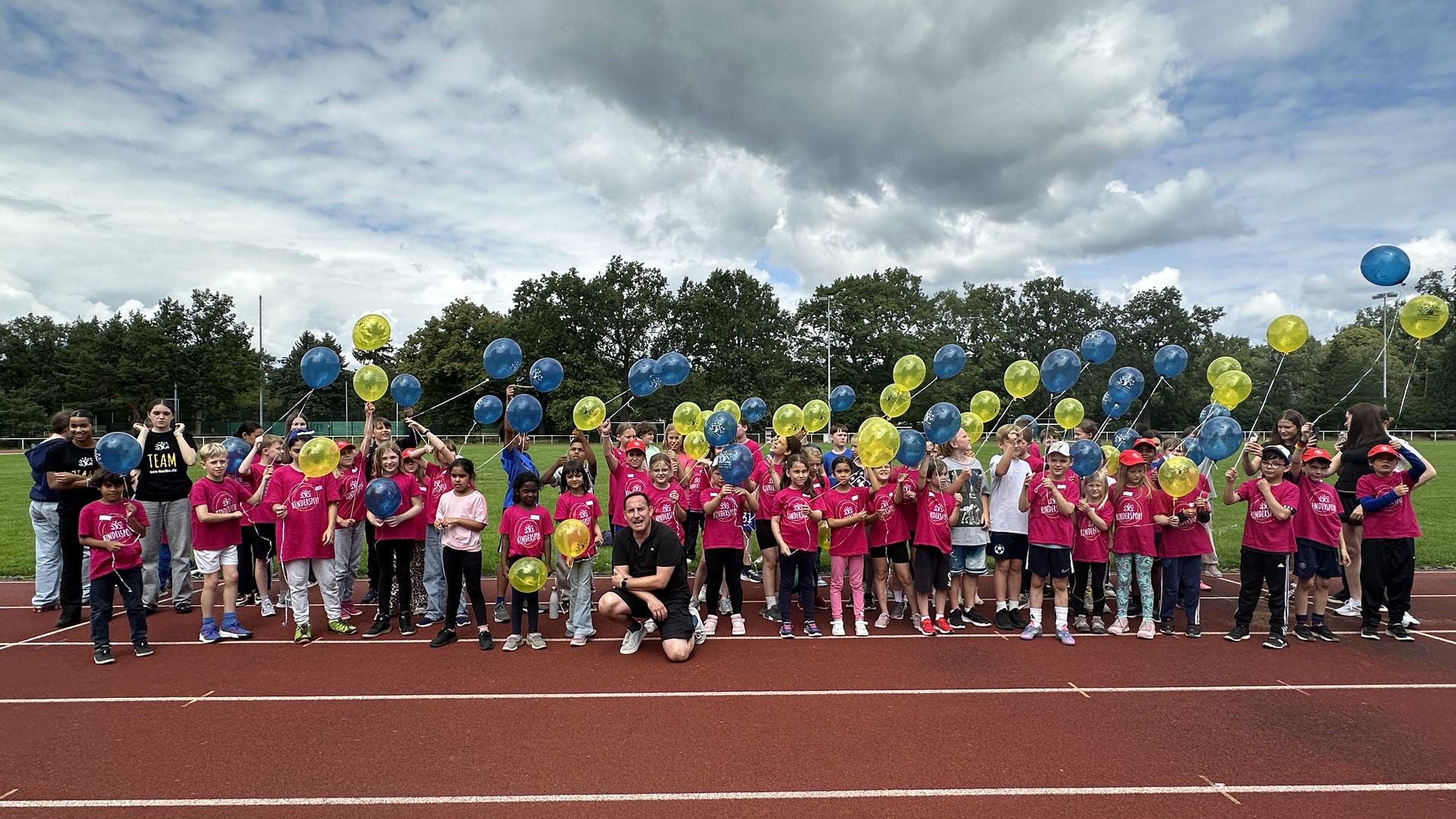 Gruppe von Kindern in pinken T-Shirts mit bunten Ballons auf einer Laufbahn, unter wolkigem Himmel.