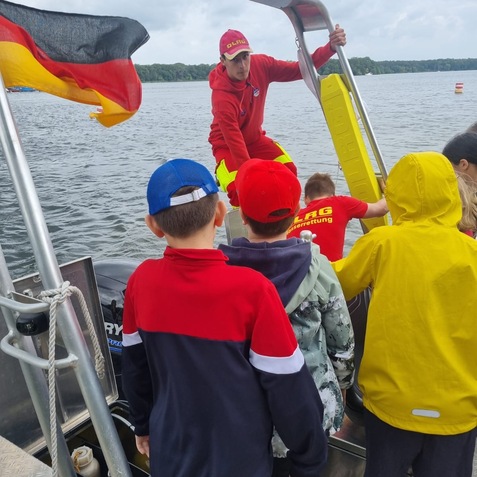 Gruppen von Kindern beobachten einen Rettungsschwimmer auf einem Boot am Wasser, im Hintergrund eine deutsche Flagge.