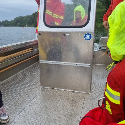 Zwei Personen in Regenjacken stehen hinter einer transparenten T&uuml;r auf einem Boot, an einem Gew&auml;sser.