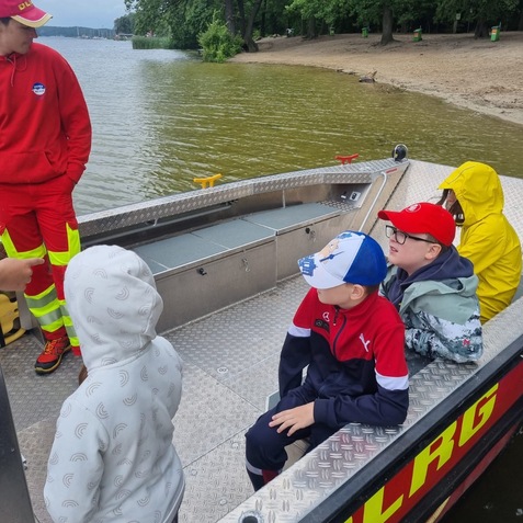 Kinder sitzen auf einem DLRG-Boot am Ufer eines Sees, Blick auf Wasser und Strand. Ein Betreuer spricht mit ihnen.