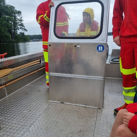 Zwei Personen in rot-gelben Uniformen stehen an einer Wasserfahrzeug-T&uuml;r, w&auml;hrend ein weiterer in gelber Regenjacke sitzt.