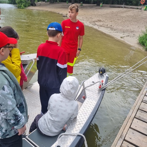 Ein Jugendlicher in rotem Rettungsschwimmer-Outfit erkl&auml;rt einer Gruppe Kinder am Ufer eines Gew&auml;ssers.