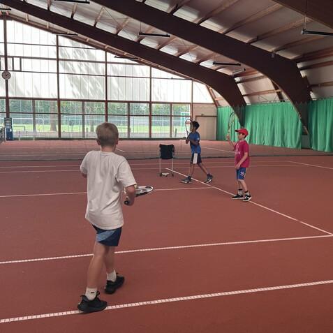 Drei Kinder trainieren Tennis auf einem roten Platz in einer Sporthalle, mit Schlägern und einem Ball.
