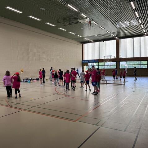 Gruppenspiel von Kindern in roten T-Shirts in einer Sporthalle mit Basketballkorb und großen Fenstern.