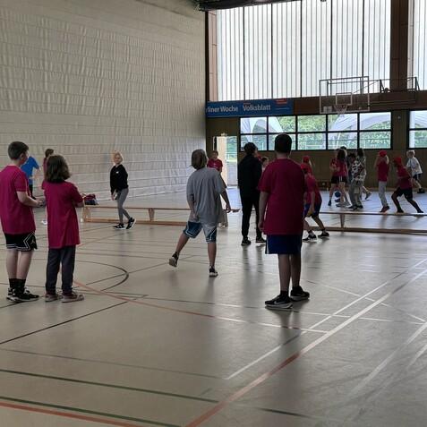 Mehrere Kinder mit roten T-Shirts spielen in einer Sporthalle, während Lehrkräfte Anweisungen geben.