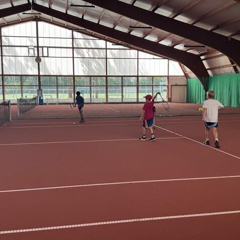 Drei Kinder spielen Tennis auf einem roten Platz in einer Sporthalle, mit Netz und großen Fenstern im Hintergrund.
