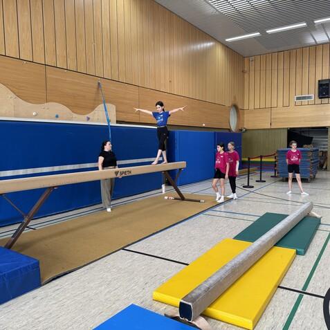 Junge turnt auf einem Schwebebalken, während Trainerin zuschaut. Weitere junge Turnerinnen stehen im Hintergrund.