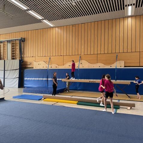 Gruppe von Turnerinnen in einer Turnhalle übt an einem Balken und auf Matten mit verschiedenen Trainingsgeräten.